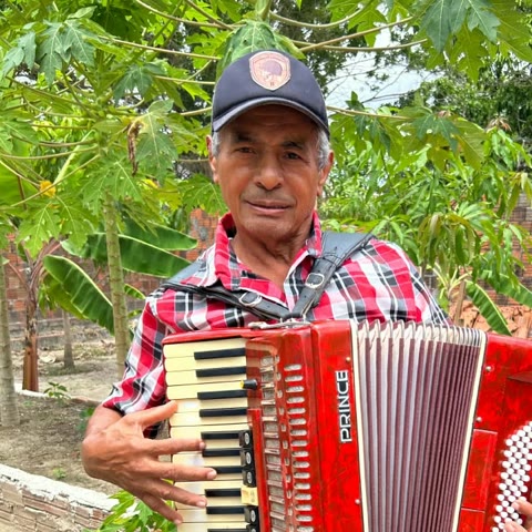 Hermínio de boné e camisa xadrez, com sua sanfona vermelha no quintal